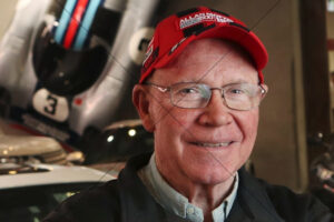 Allan Moffat smiling with race cars behind 6" x 4" Photo Print
